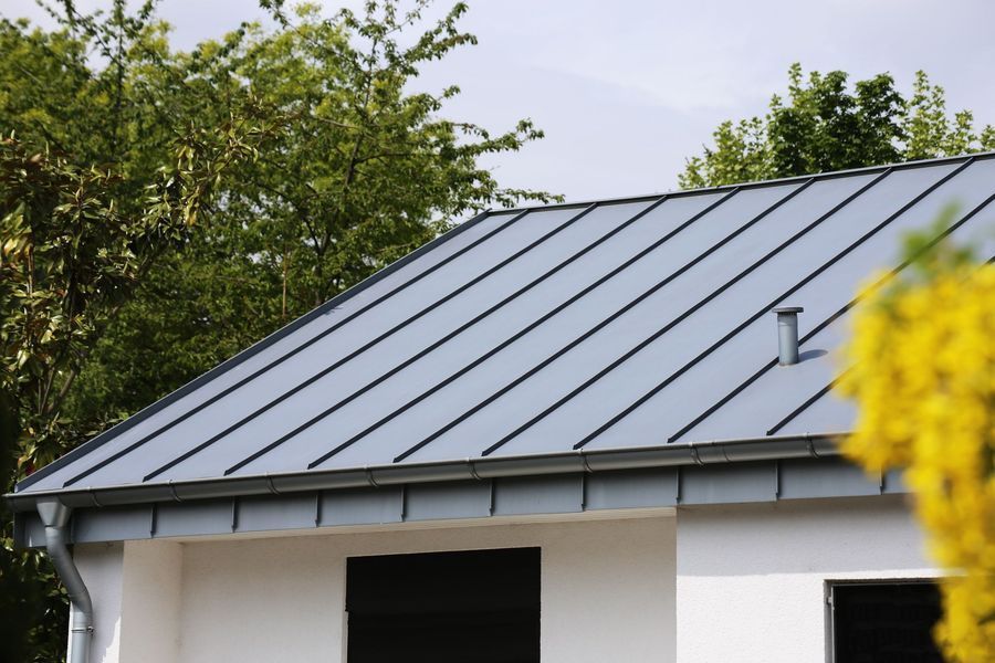 Gray metal roof on a white house, with a gutter and trees in the background.