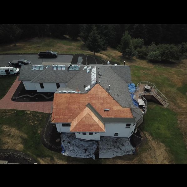 Overhead view of a house undergoing roof replacement. Crews on roof, tarps on ground, cars parked nearby.