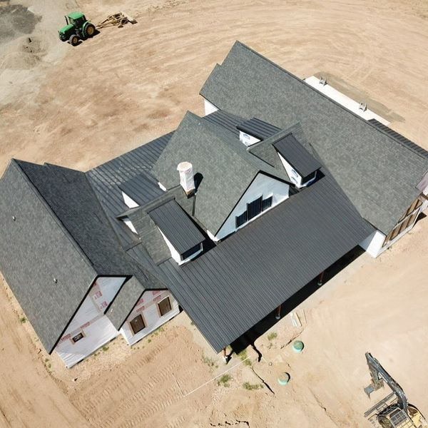 Aerial view of a house under construction with dark gray roof and white siding; a tractor and excavator in the dirt.