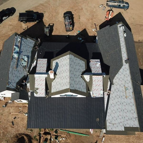 Aerial view of a house under construction with different roof sections, some covered in dark grey shingles.