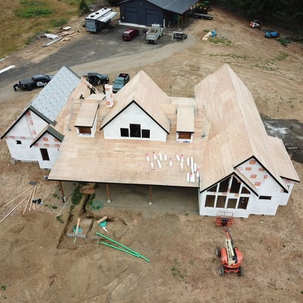 Construction site with a new house; roof being installed. White siding, wood roof. Vehicles and equipment visible.