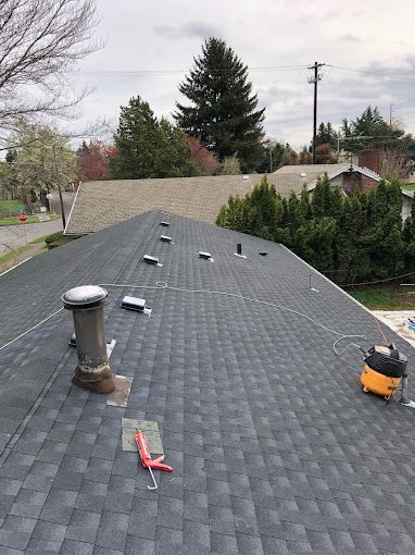 Roof with dark shingles and vent pipes. A vacuum and tools sit on the roof. In background, a neighborhood with trees and homes.