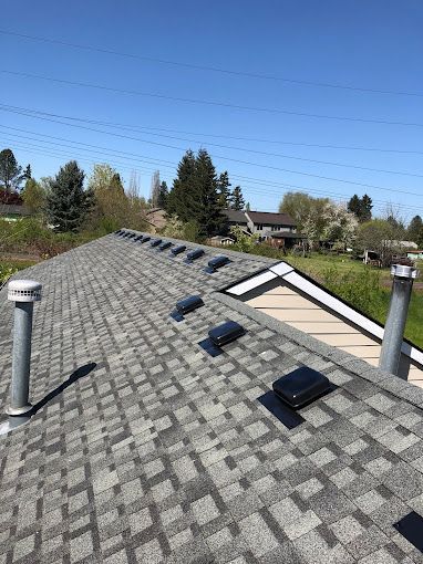 A roof with gray asphalt shingles, multiple vents, and chimneys under a blue sky.