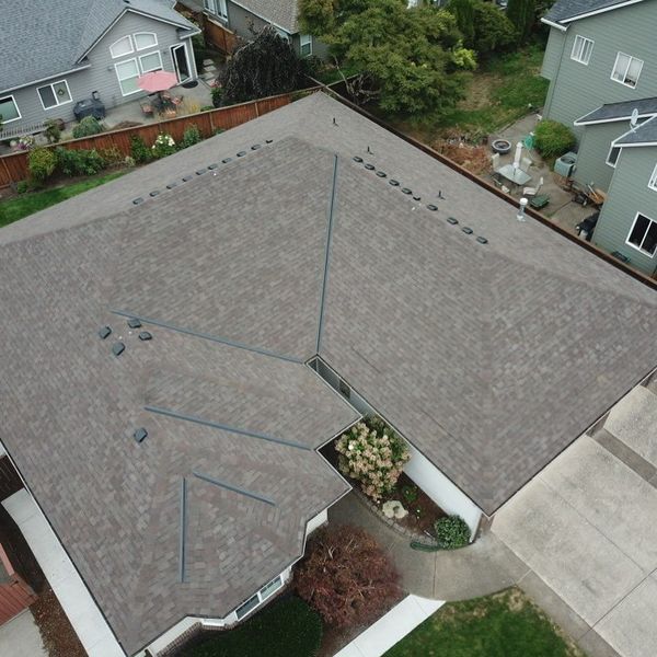 Overhead view of a house with a brown shingle roof, surrounded by other houses and green trees.