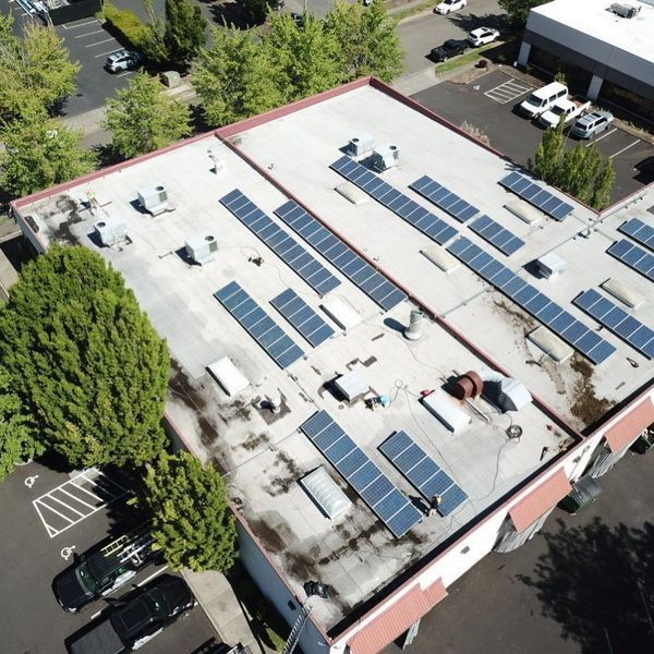 Aerial view of a commercial building with solar panels on its flat roof. Parking lot and trees surround it.