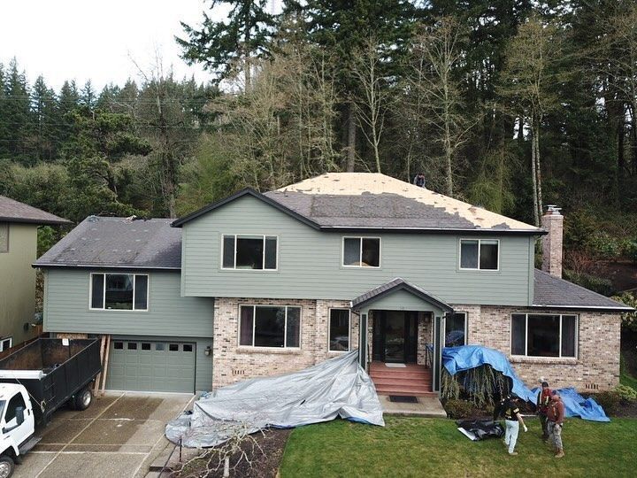 House with a partly-shingled roof and construction materials, green siding, brick facade, trees in the background.