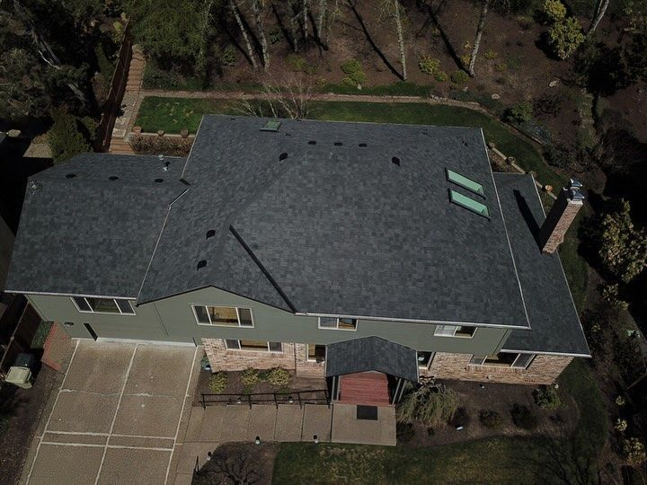 Overhead view of a two-story house with a dark gray shingle roof, surrounded by trees and a concrete driveway.