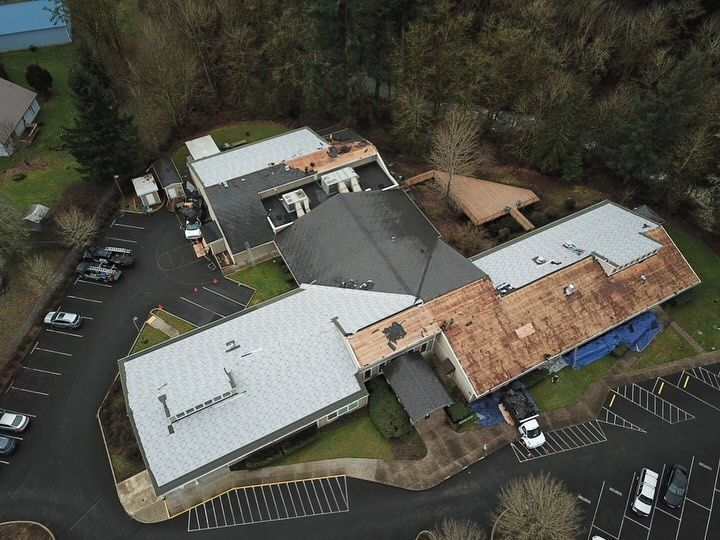 Aerial view of a complex with multiple gray and brown roofs, a parking lot, and surrounding trees.