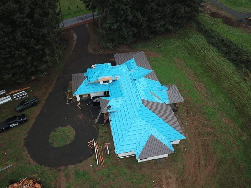 Aerial view of a house under construction with blue roof underlayment and gray roofing. Black asphalt driveway.