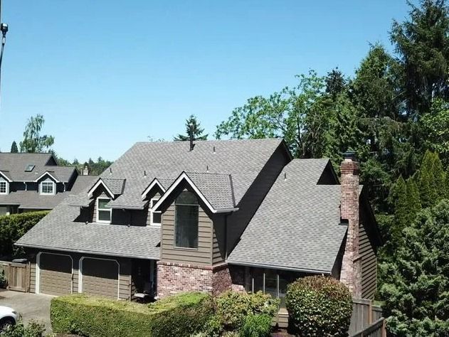 House with brown siding, brick, and gray shingle roof, surrounded by green trees and blue sky.