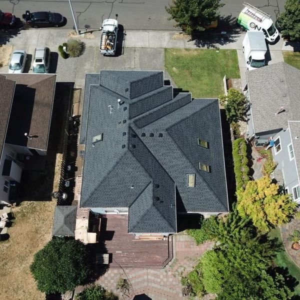 Aerial view of a house with a complex, dark gray roof, surrounded by cars, trees, and other buildings.