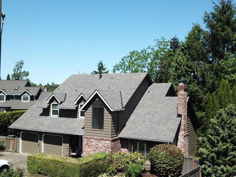 House with gray shingle roof, brown siding, brick chimney, three dormers, and a garage, surrounded by trees.