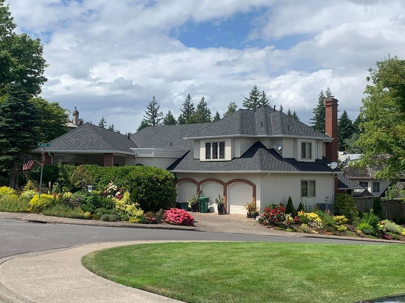 White house with black roof, three-car garage, and well-maintained landscaping under a cloudy blue sky.