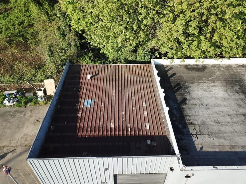Overhead view of a warehouse roof. One section is corrugated brown metal, another is black. Green trees in background.