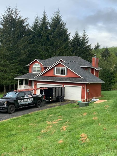 Red house with a dark gray roof. Black truck and trailer in the driveway. Green lawn, trees in the background.