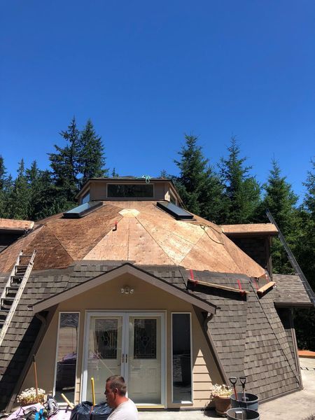 Dome-shaped house under construction with brown roof shingles. A man works near the entrance on a sunny day.