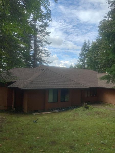 Brown-roofed house with windows, surrounded by trees and a grassy yard under a cloudy sky.