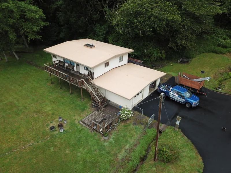 Elevated house with beige roof and siding, brown deck, driveway, surrounded by greenery. A blue vehicle is parked in front.