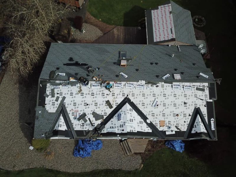 Overhead view of a house roof partially under construction. Workers are replacing shingles, with materials and tarps visible.