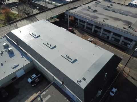 Aerial view of a gray industrial building with vents on the roof. Adjacent buildings and street visible.
