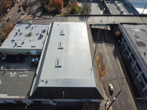 Aerial view of industrial buildings with gray roofs, street, and parked cars.
