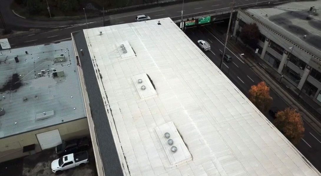 Overhead view of a long, flat commercial building roof with vents. A road and other buildings are in the background.