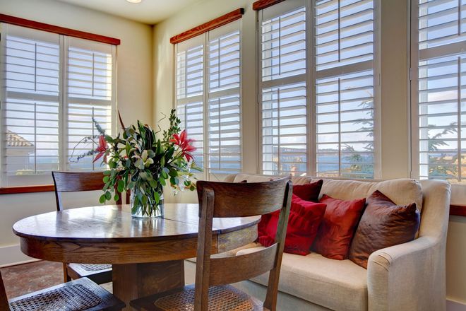 Bright dining nook with round table, flowers, beige sofa, and large windows with white shutters.