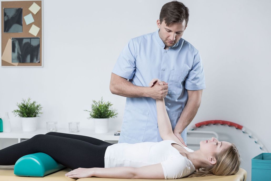 Physical therapist stretches patient's arm; patient lies on a therapy table, in a clinic setting.