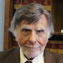 Older man with gray hair and beard, in suit, looks at camera.