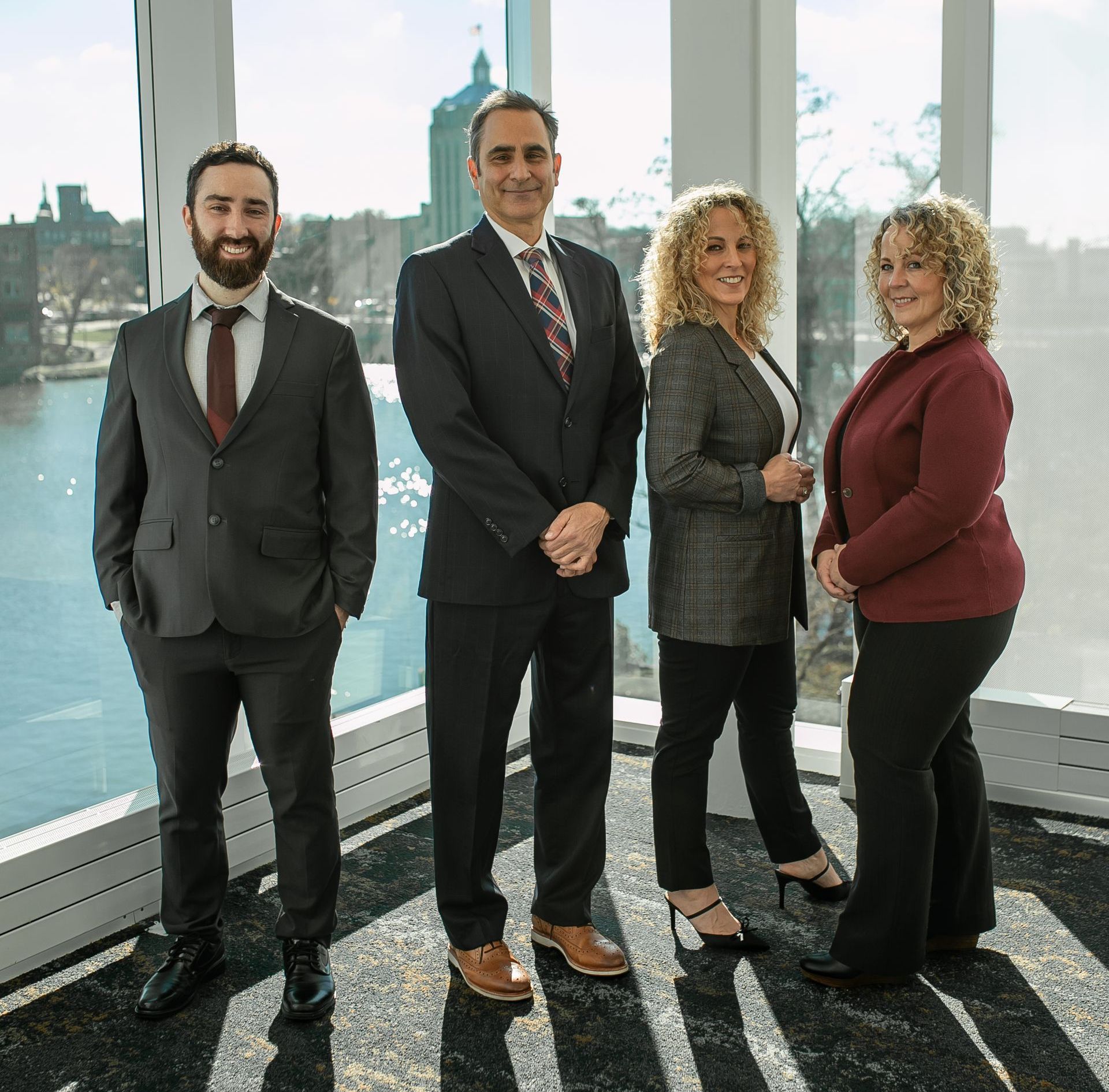 Four professionals in suits pose by a large window overlooking a city and river.