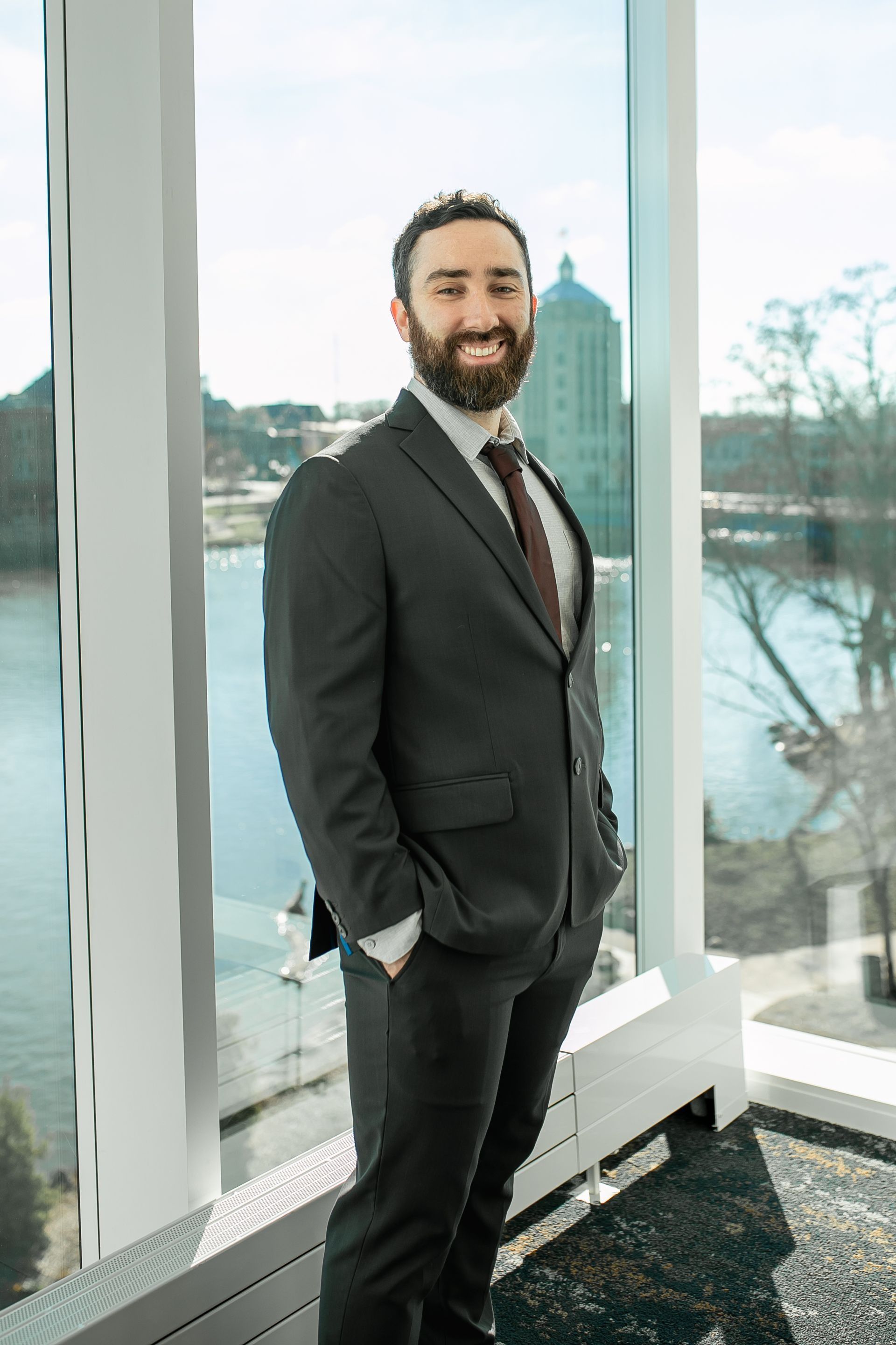 Man with beard in suit, hands in pockets, smiling near a window with river view.