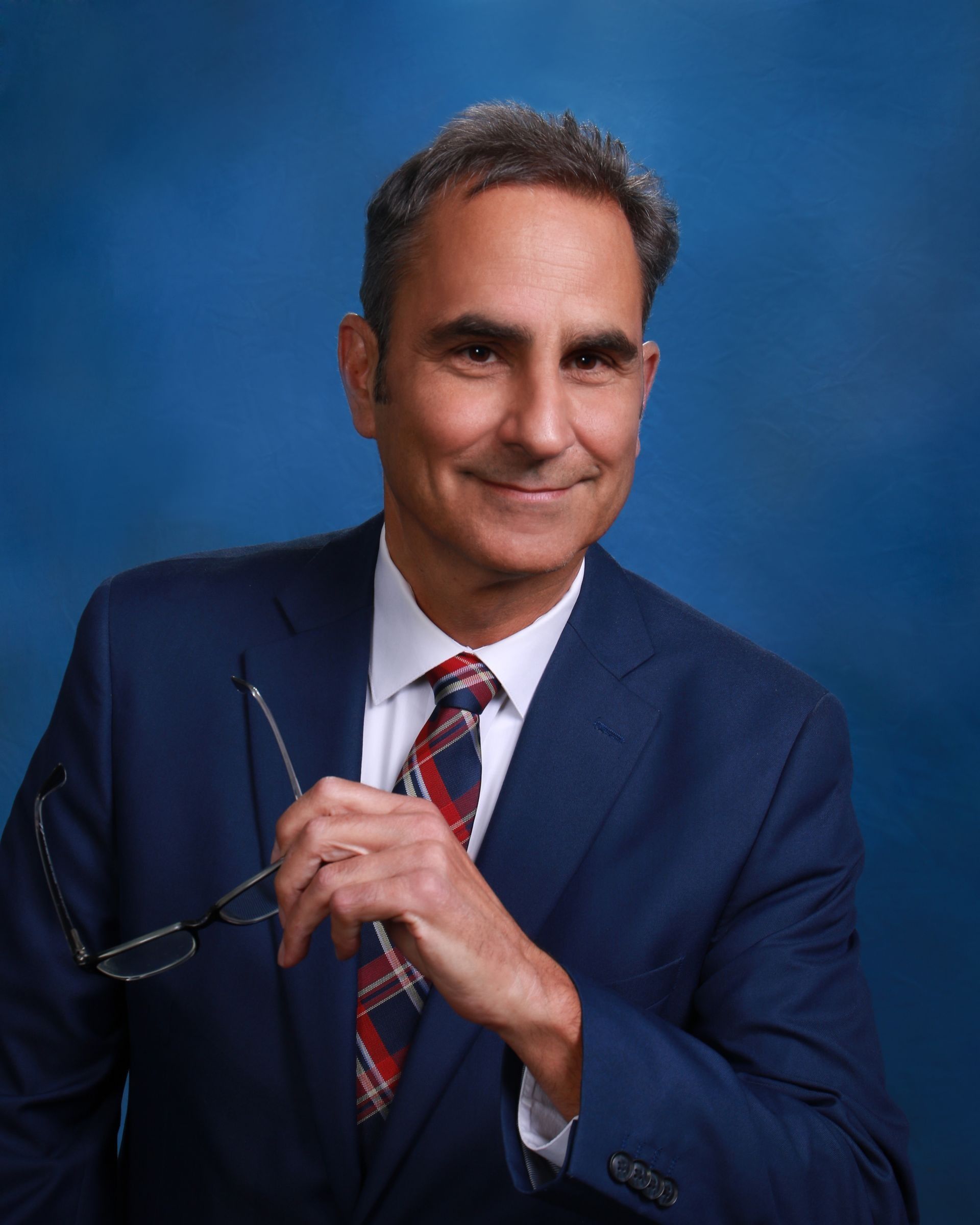 Man in suit, holding glasses, smiling. Blue backdrop.