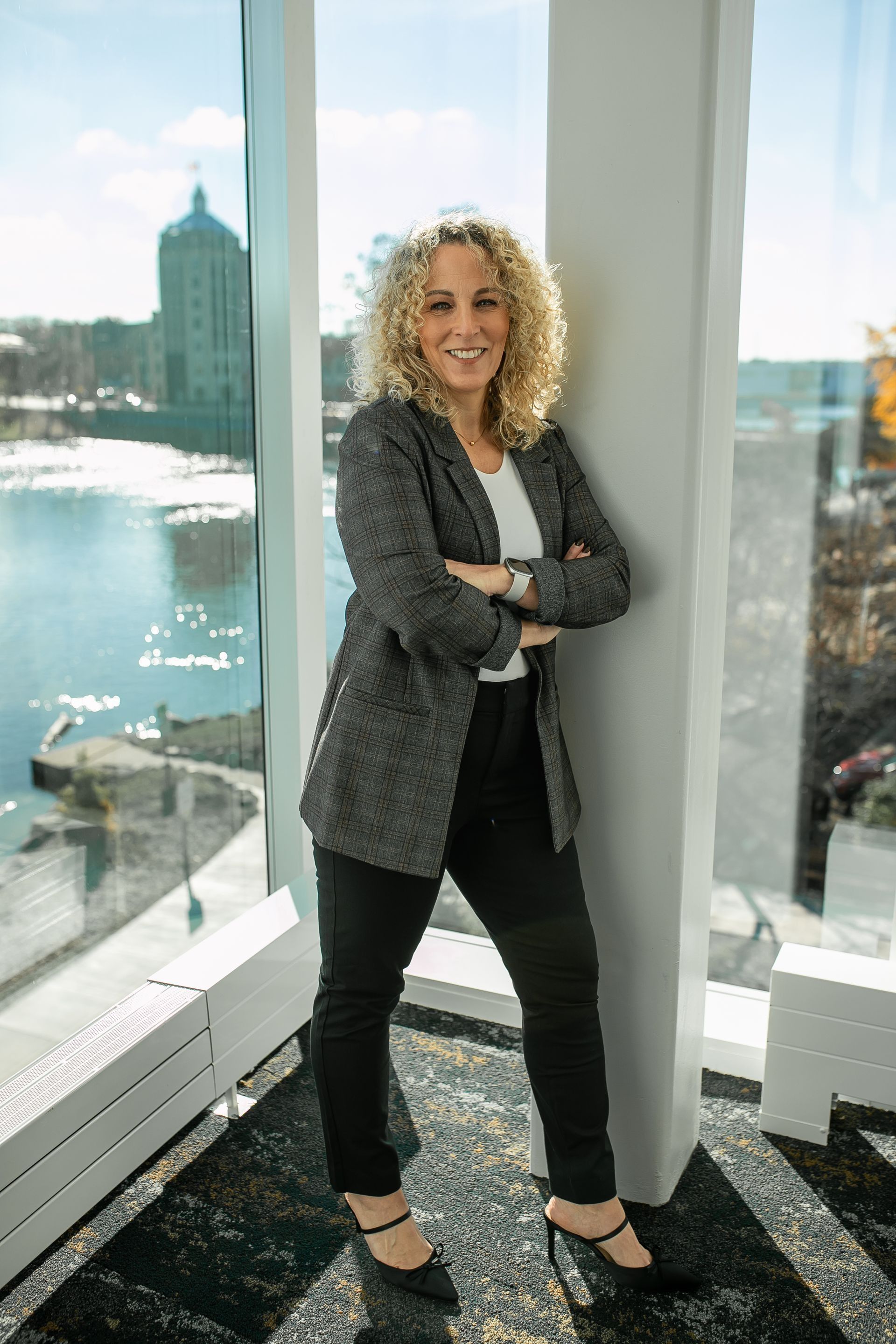 Woman with curly blonde hair leans against a white pillar, arms crossed, smiling. Window shows a cityscape.