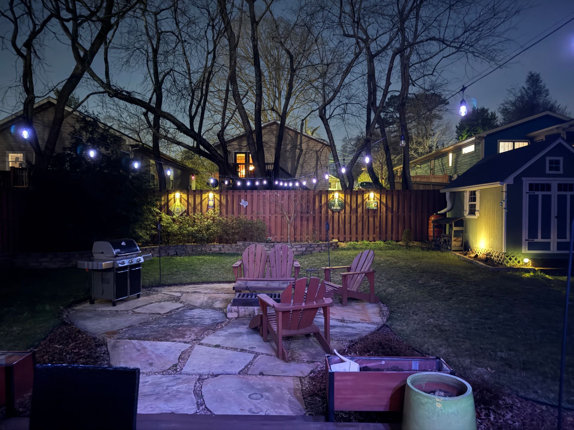 A backyard patio at dusk with wooden chairs, a grill, and string lights illuminating a stone patio and fenced garden.