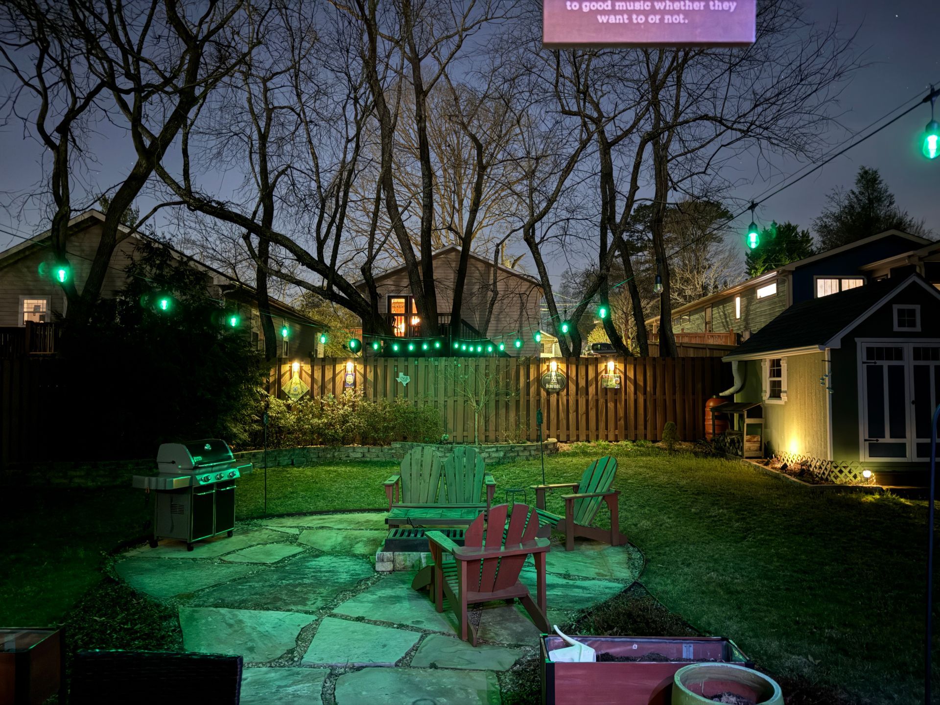 A twilight backyard scene features a stone patio with outdoor chairs, a fire pit, and glowing green string lights.