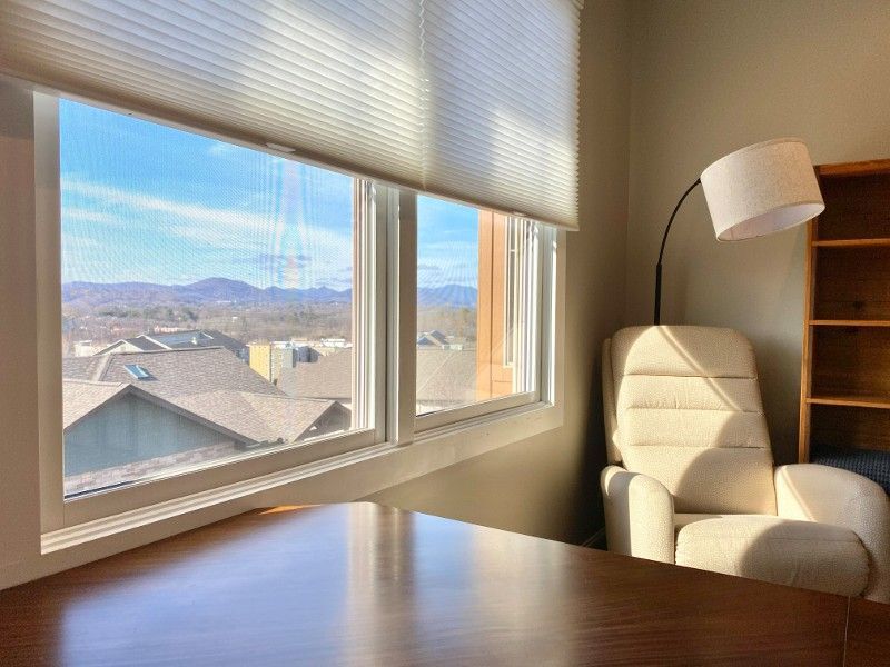 A room with a desk , chair , lamp and window with a view of a city.