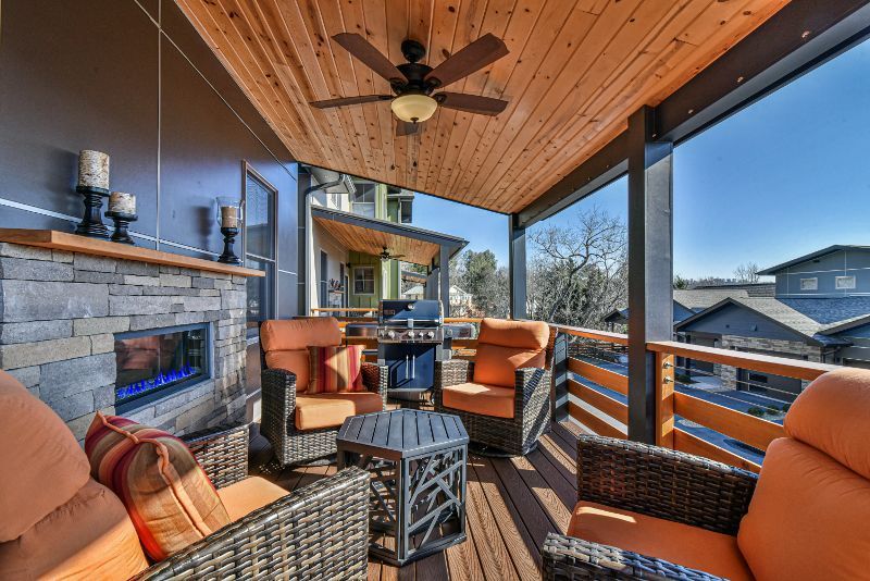 A porch with a fireplace , chairs , a table and a ceiling fan.