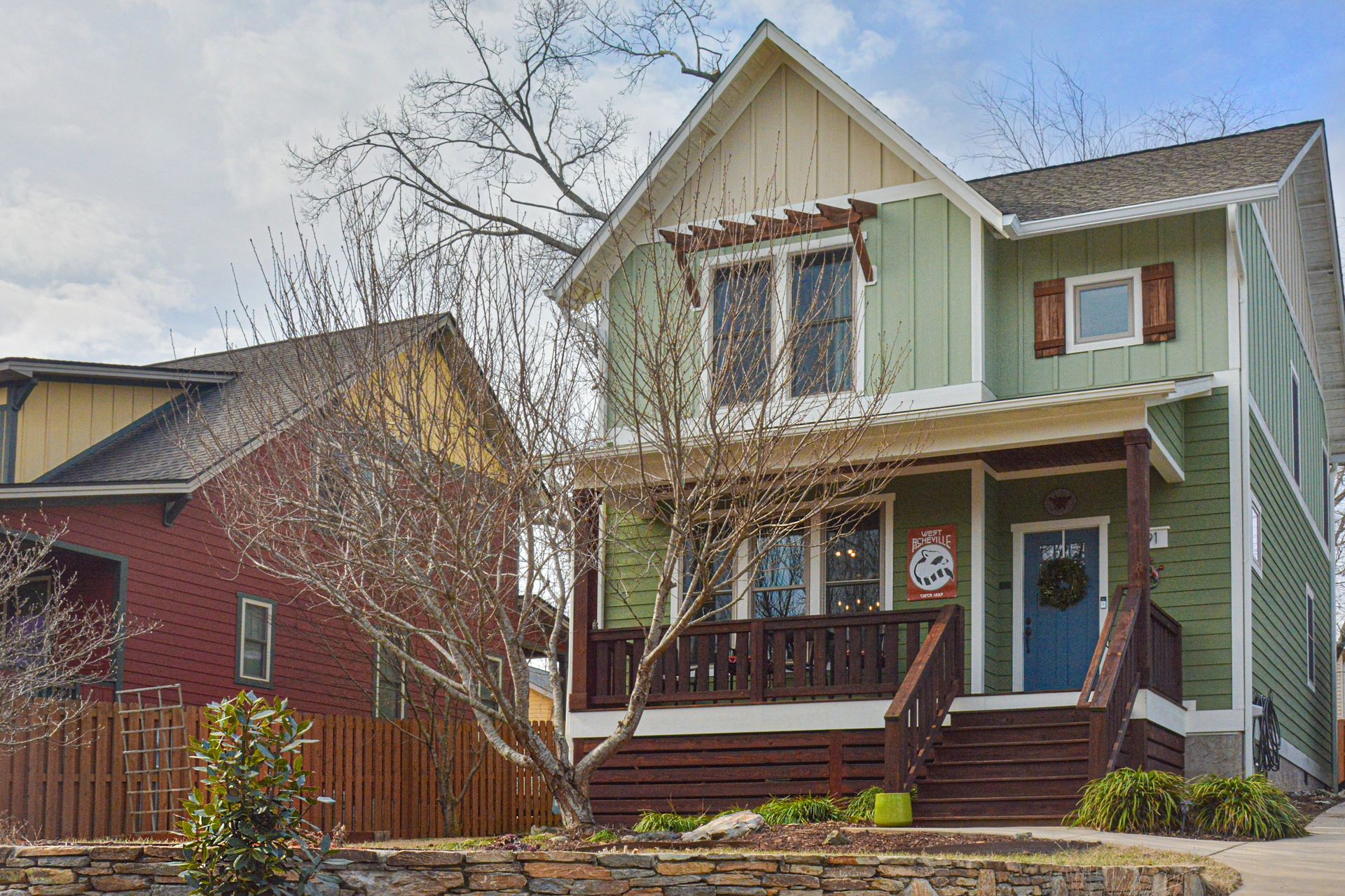 A sage green two-story house with a blue front door, porch, and brown trim, next to a red house with a wooden fence.