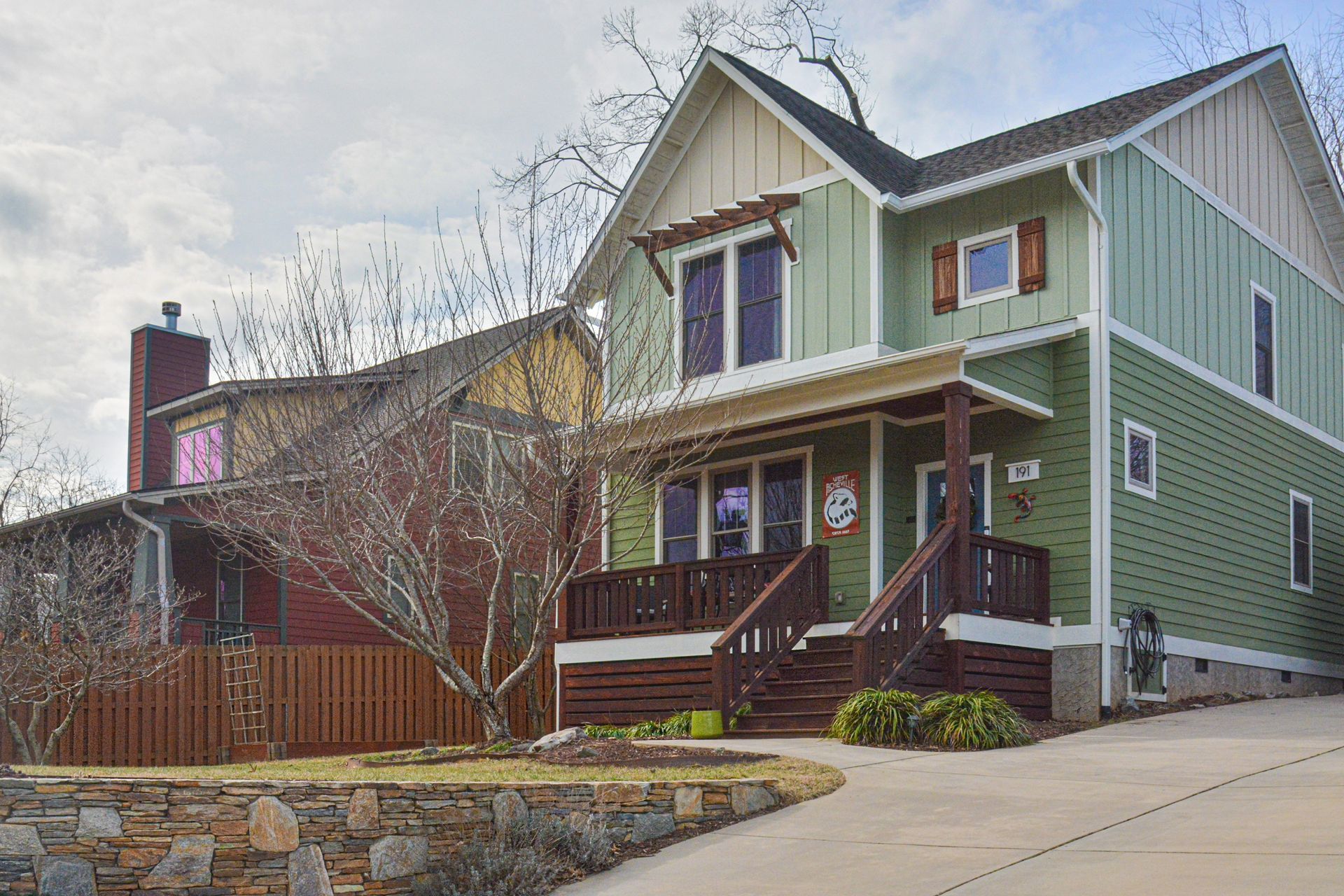 A two-story green house with a front porch and a wooden fence, next to a brown house with a stone foundation.