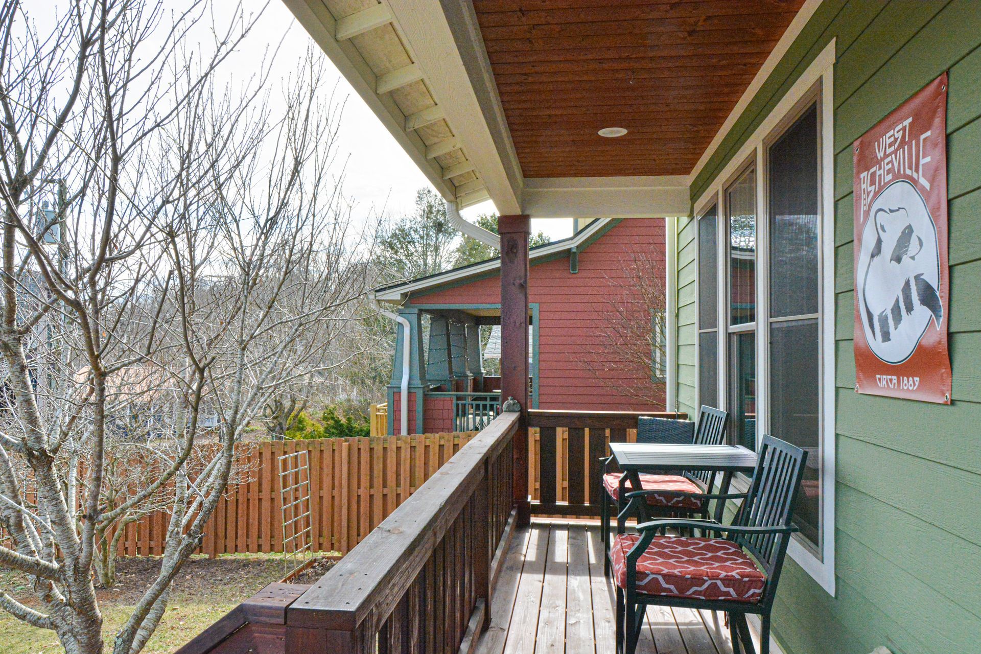 A green wooden porch featuring a seating area with chairs and a patterned red banner on the wall, overlooking a yard.