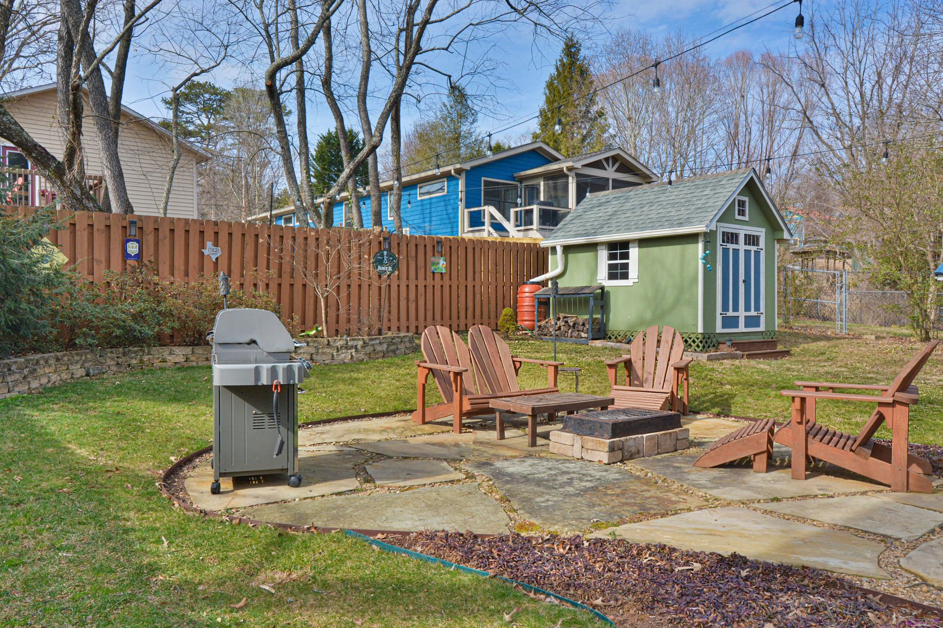 A backyard patio with a grill, fire pit, and wooden chairs next to a small green shed and a tall wooden fence.