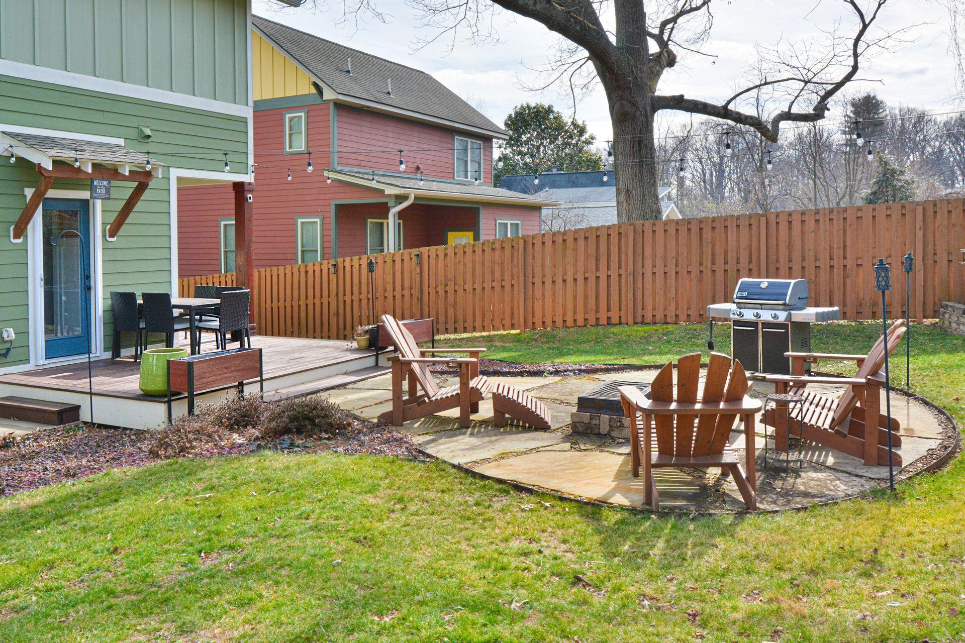 Backyard patio with wooden chairs, a fire pit, and a grill, set against a wooden fence and neighboring houses.