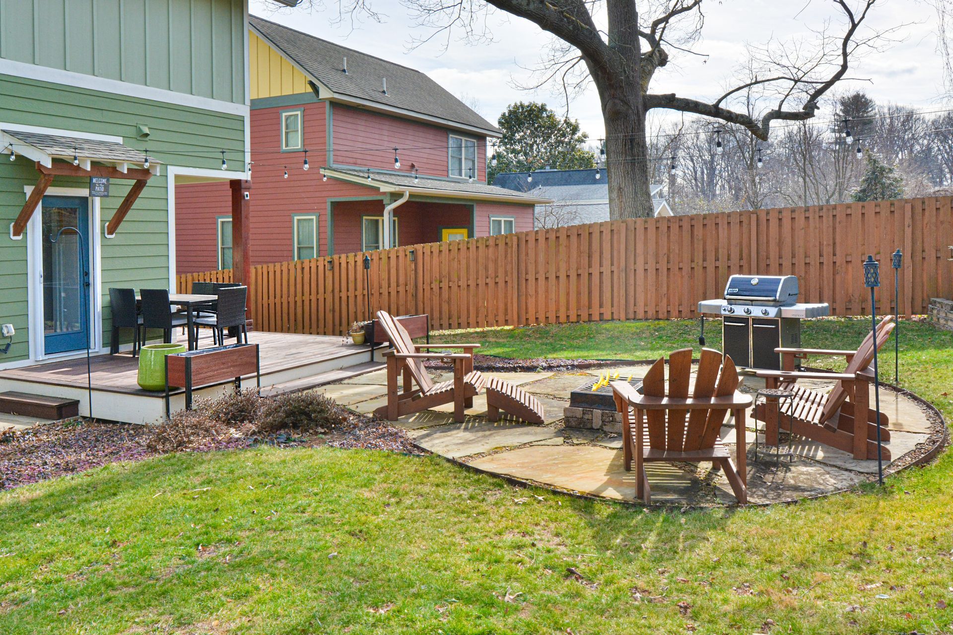 A stone patio in a backyard features wooden Adirondack chairs arranged around a fire pit near a grill and a fence.