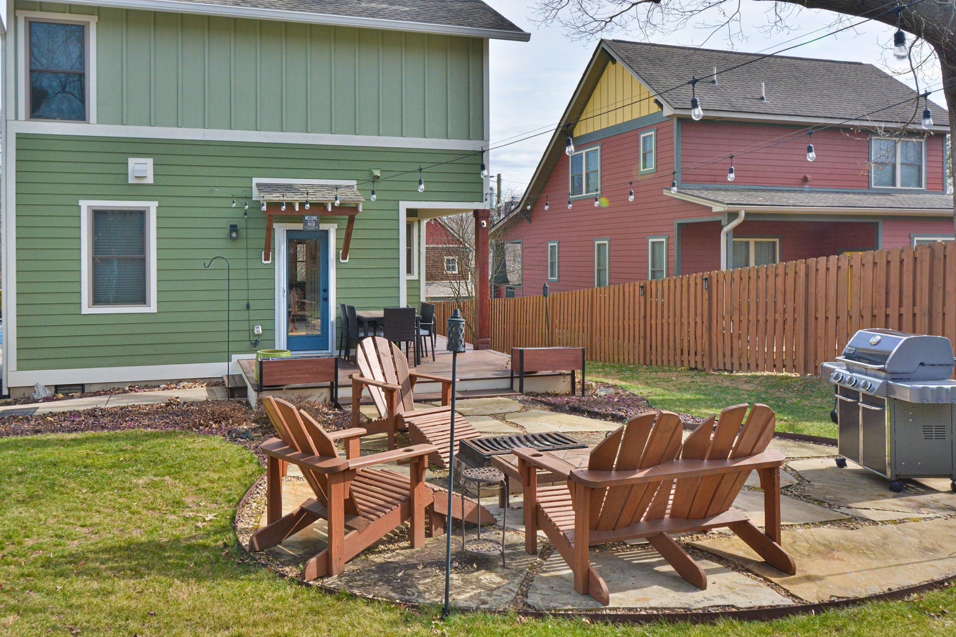 A green house backyard with a stone patio, wooden Adirondack chairs, a fire pit, and a grill in front of a wooden fence.