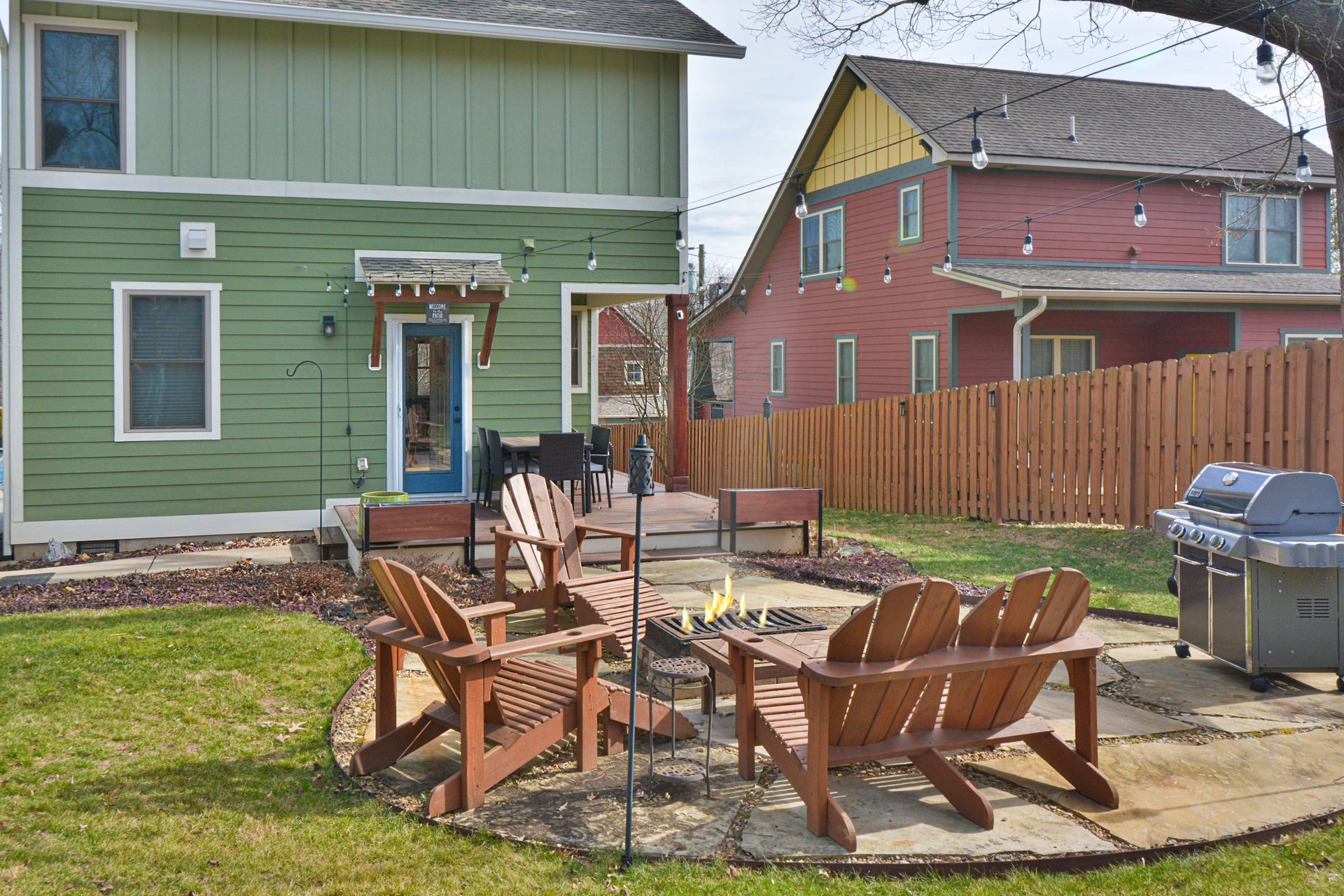 A backyard patio with a fire pit, brown wooden chairs, and a grill, set between a green and a red house.