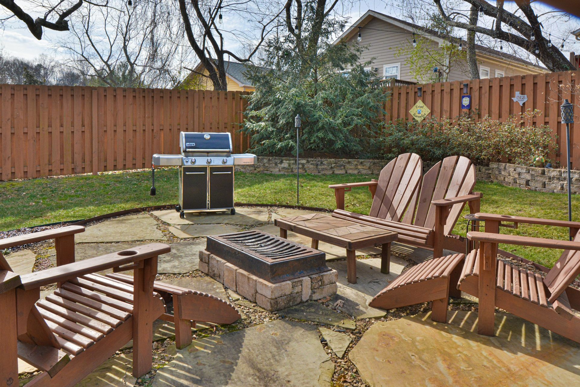 A backyard patio with a fire pit, wooden chairs, and a grill, set against a wooden fence and grassy lawn.