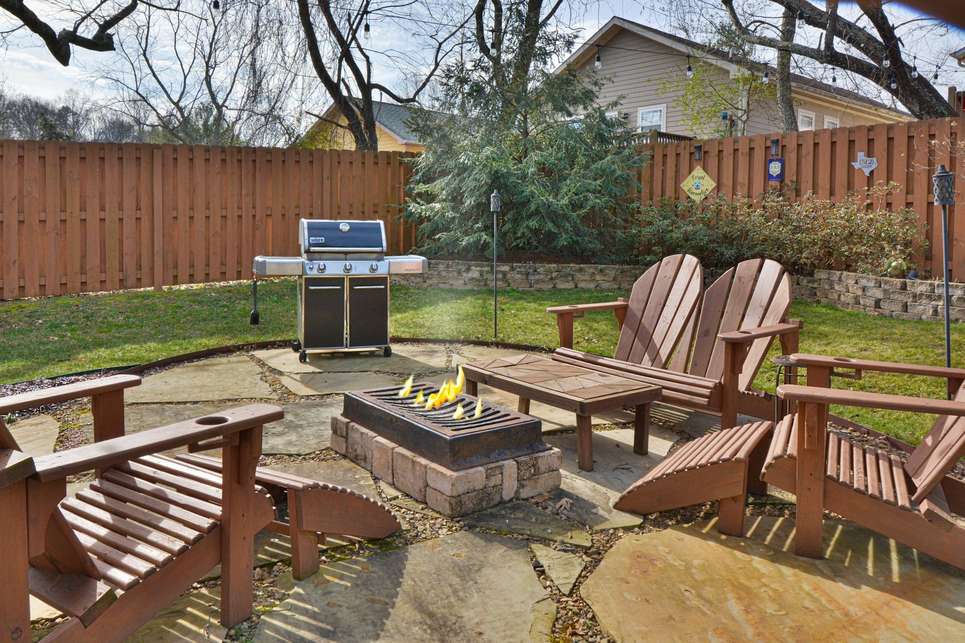 A backyard stone patio with a lit fire pit, three wooden Adirondack chairs, and a gas grill in front of a wooden fence.