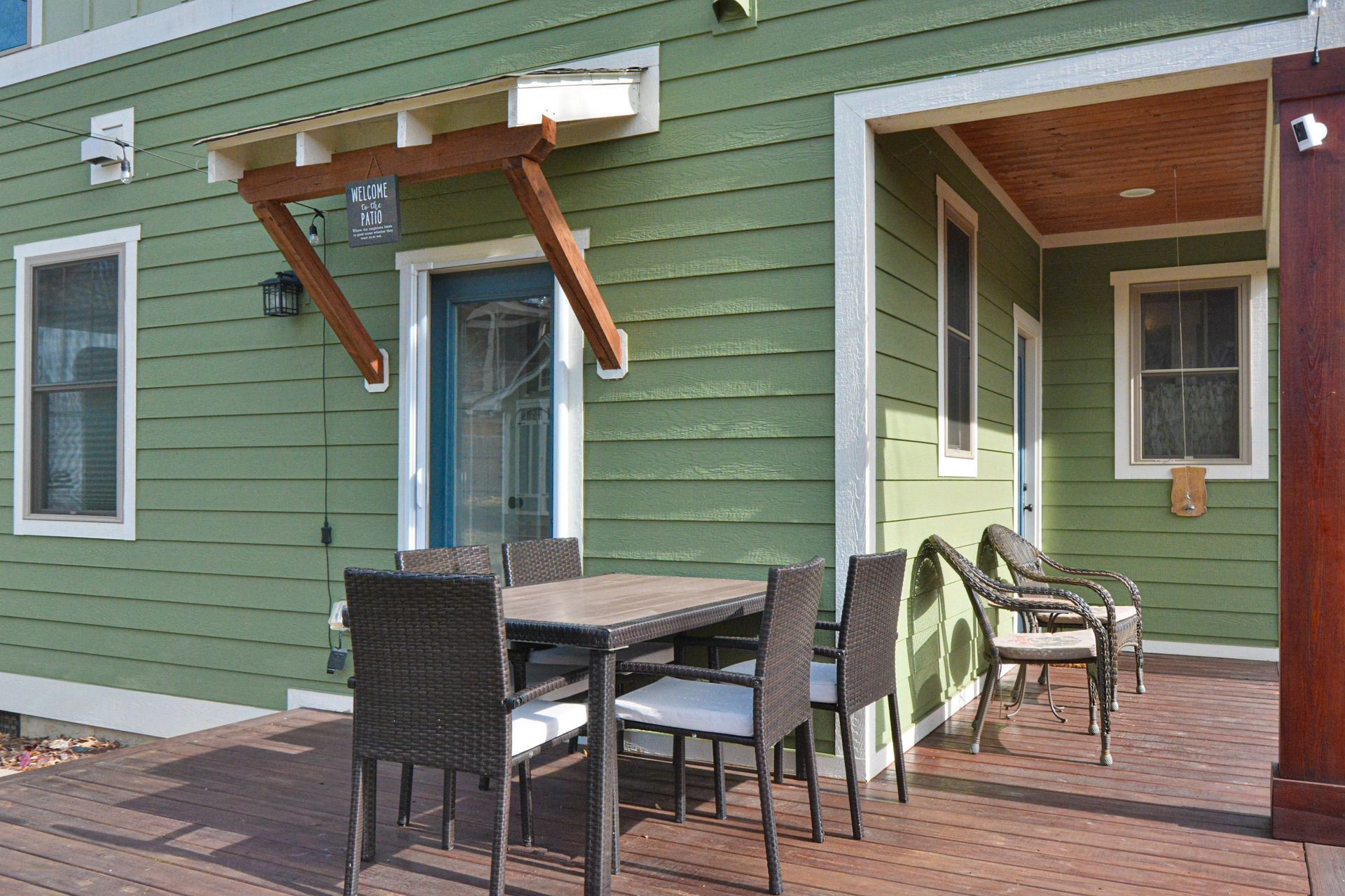 A green-sided house features a covered patio with an outdoor dining set and chairs on a paved deck.