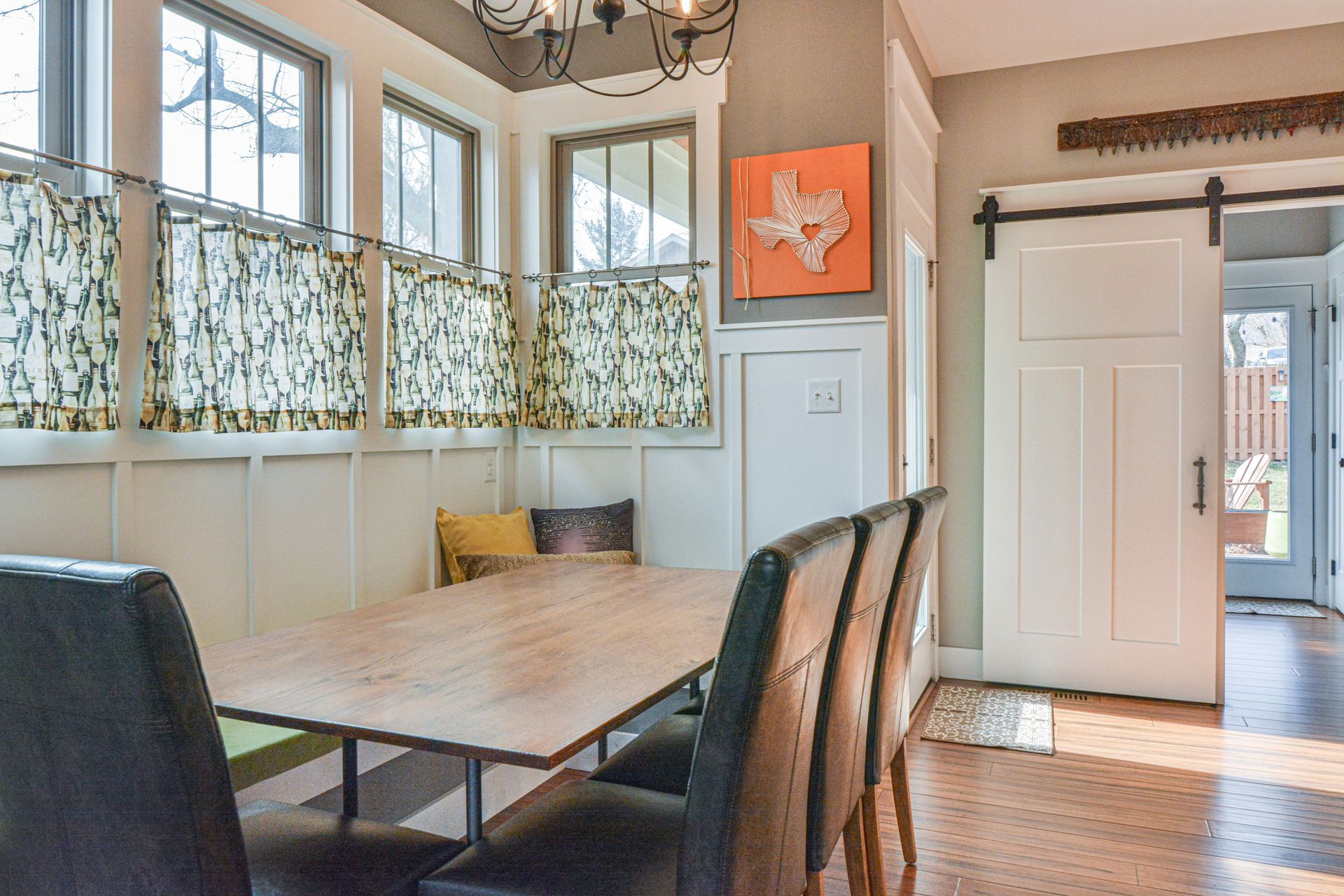 A dining area with a wooden table, dark chairs, a built-in bench under windows with patterned curtains, and a sliding door.