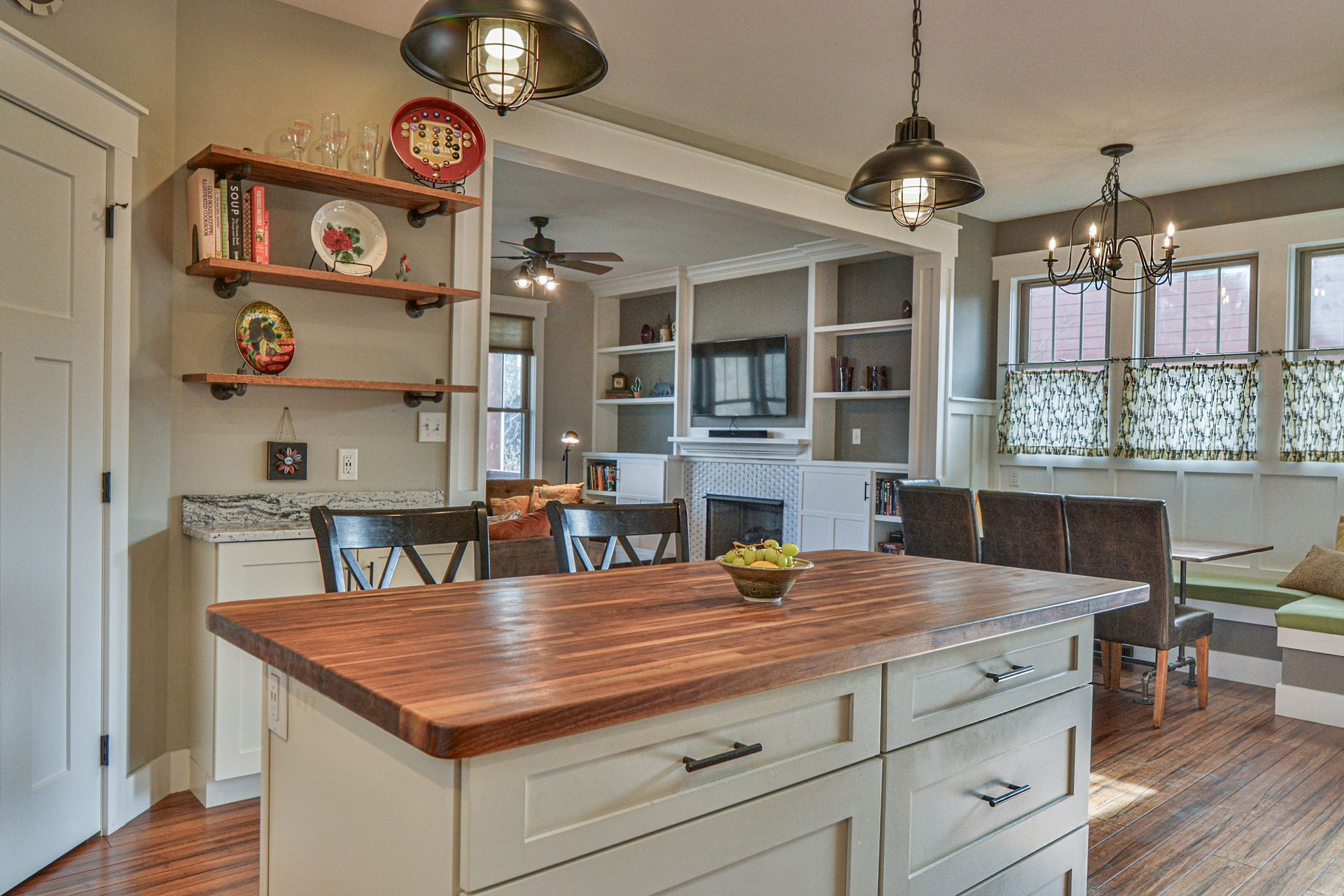 A kitchen with a wooden island, grey cabinets, open shelving, and a dining nook with a bench, viewed from the living area.